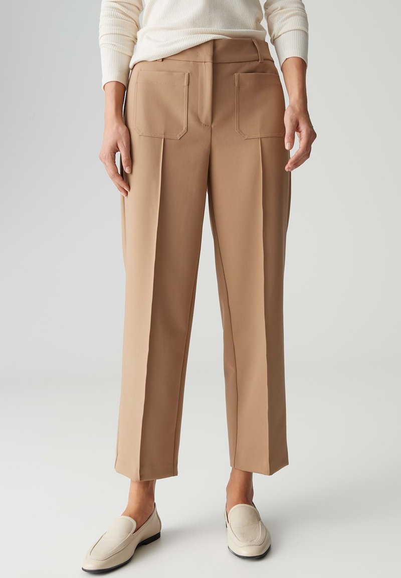 Beige geplooide broek met een rechte pijp, voorzien van twee achterzakken en een gladde textuur. Gecombineerd met beige instapschoenen.