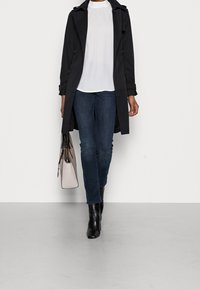 Trench-coat noir sur un chemisier blanc, associé à un jean bleu foncé et des bottines noires. Un sac à main rose clair est tenu dans la main droite.