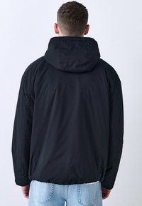 Hombre con chaqueta con capucha negra y jeans de color azul claro, visto desde la espalda contra un fondo liso claro.