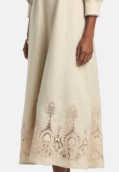 Beige linnenjurk met ingewikkeld kanten borduurwerk langs de zoom, gedragen door een persoon met een donkere huid en een gouden armband om één pols.