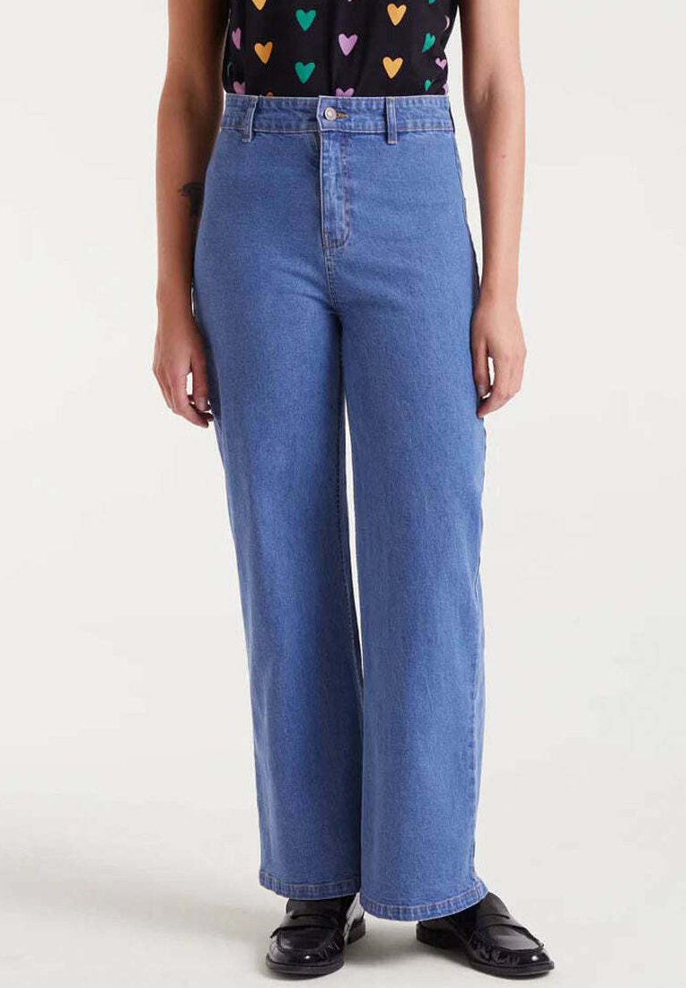 Mujer vestida con jeans azules de tiro alto y pierna recta, mocasines negros y una blusa negra con patrones de corazones coloridos.