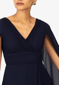 Abito blu navy con scollo a V, caratterizzato da dettagli plissettati sul corpino e maniche a mantello fluttuanti, realizzato in un tessuto leggero e morbido.