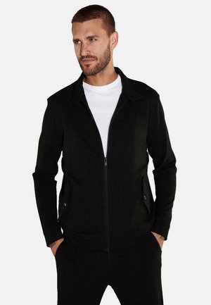 Black zip-up jacket with a collar, two front pockets, and a textured fabric. Worn over a white t-shirt, fitted design, and long sleeves.