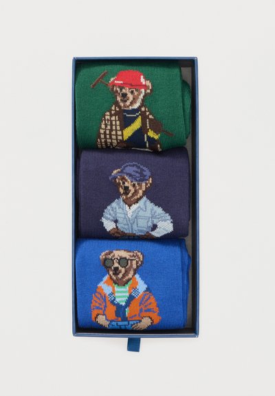 Τρία ζευγάρια διπλωμένες κάλτσες σε ένα κουτί, το καθένα με μια αρκουδάκι που φοράει διαφορετικές στολές και καπέλα σε πράσινο, ναυτικό και μπλε φόντο.