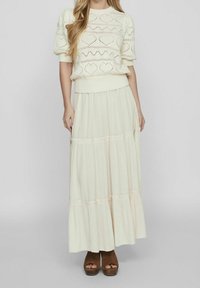 Krémový pletený svetr se vzory srdcí, v kombinaci s volnou, vrstvenou maxi sukni. Hnědé platformové boty doplňují komplet.