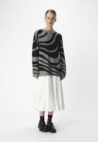 Oversized grå sweater med sorte bølgede striber, parret med en hvid nederdel. Sorte sko med spænder og gennemskinnelige sorte sokker.