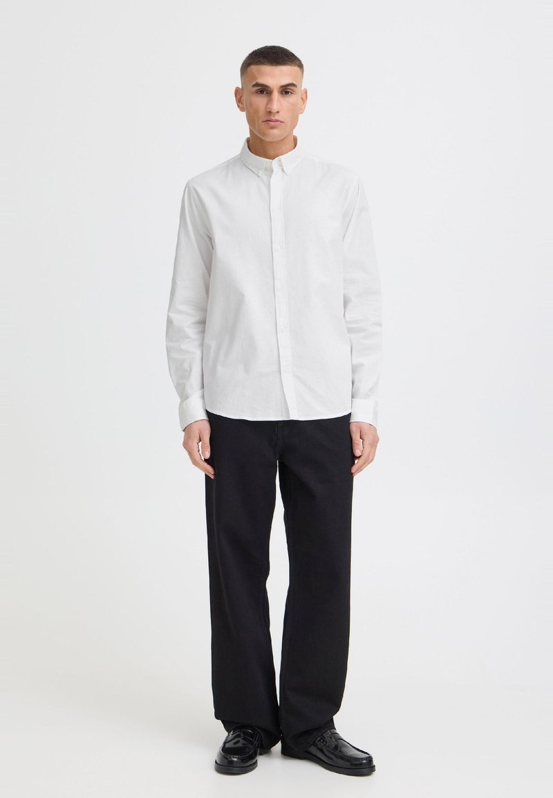 Homme debout portant une chemise blanche à manches longues boutonnée, un pantalon noir ample et des chaussures habillées noires sur un fond blanc uni.