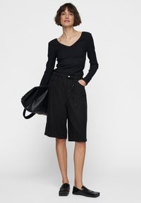 Zwarte ribbeltrui met lange mouwen, gecombineerd met losse zwarte knielange shorts. Model houdt een gestructureerde zwarte tas vast. Eenvoudig, minimalistisch ontwerp.
