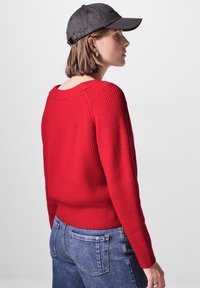 Roter gerippter Pullover mit weitem Ausschnitt und langen Ärmeln, kombiniert mit blauen Jeans. Eine graue Baseballmütze rundet das Outfit ab.