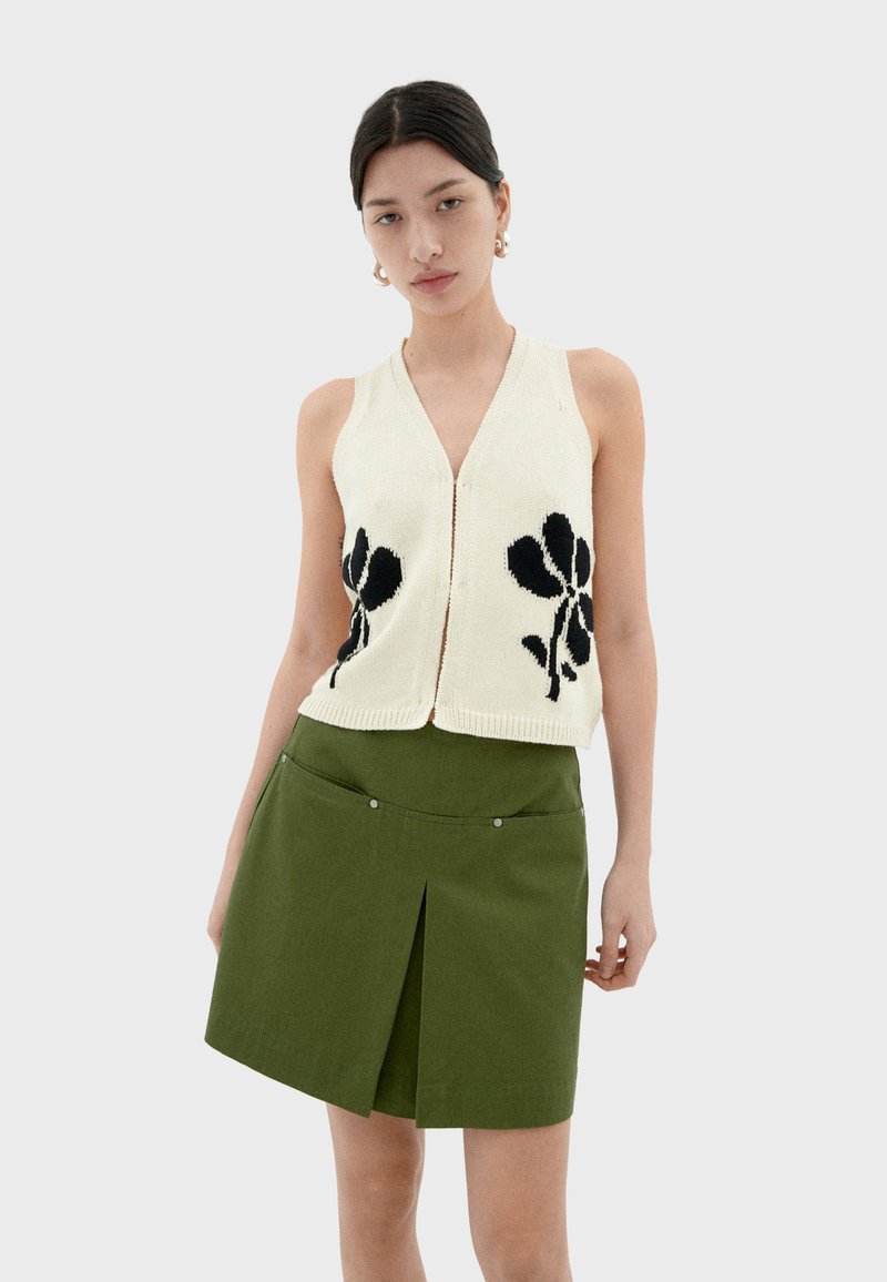 Crème gebreide vest met zwart bloemenpatroon, gecombineerd met een groene rok met een voorplooien en decoratieve hardware-accenten.