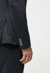 Férfi keze sötétszürke öltönyzakó ujjának oldalán, amelyen négy gomb van, és hozzá illő nadrág világos, egyszínű háttér előtt.