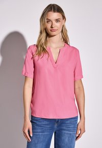 Kurzärmelige rosa Bluse mit V-Ausschnitt. Aus leichtem Stoff gefertigt, mit umgeschlagenen Ärmelaufschlägen und geradem Saum. Kombiniert mit blauen Jeans.