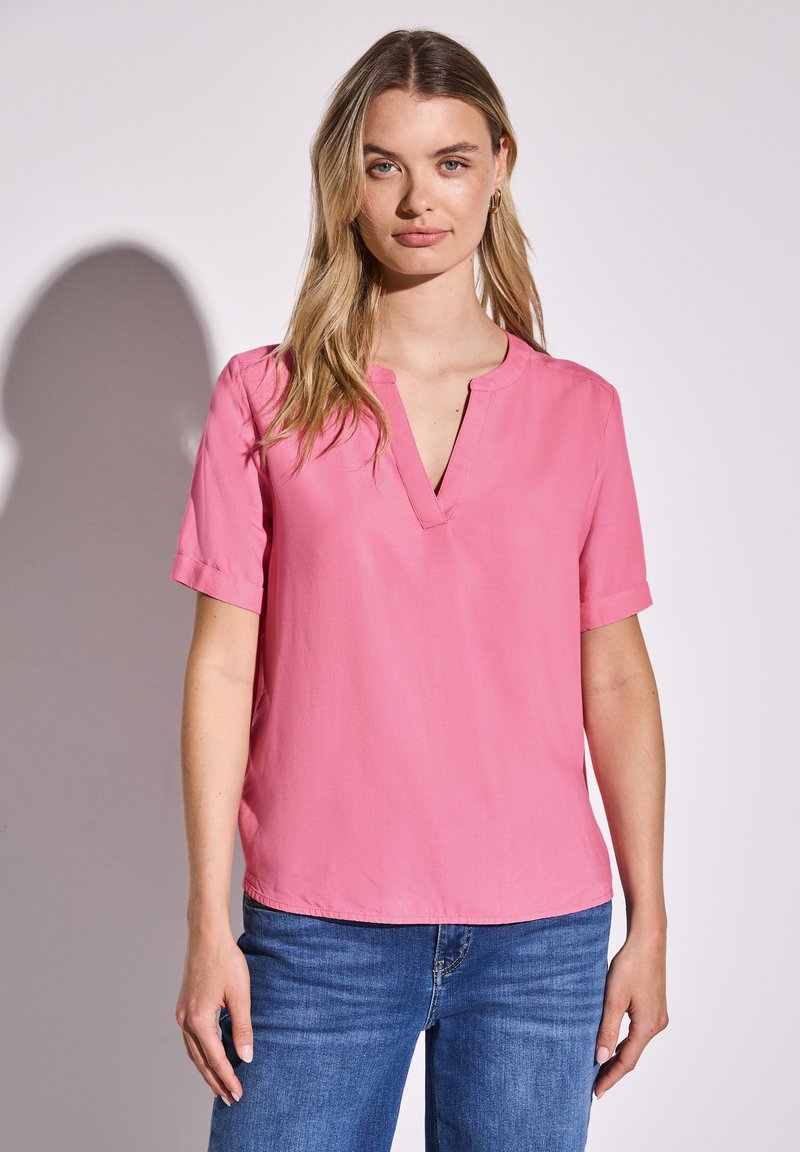 Kurzärmelige rosa Bluse mit V-Ausschnitt. Aus leichtem Stoff gefertigt, mit umgeschlagenen Ärmelaufschlägen und geradem Saum. Kombiniert mit blauen Jeans.