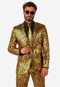 Zelta un melnas krāsas uzvalks ar zvaigžņu rakstiem, izceļas ar vienu pogu aizdari, šaura piegriezuma un saskaņotu kaklasaiti. Kombinēts ar tumšām saulesbrillēm.