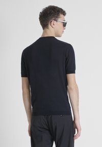 Maglione corto a maniche corte di colore nero con scollo rotondo e orlo a coste. Tessuto liscio e design aderente. Indossato con pantaloni scuri e occhiali da sole.