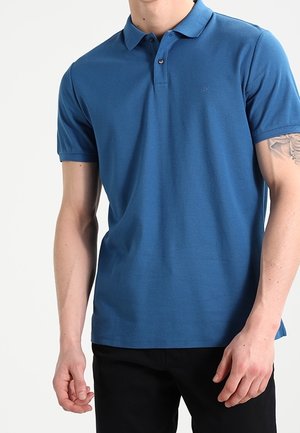 Mand iført en blå kortærmet polo og sorte bukser, med en synlig tatovering på hans venstre overarm.