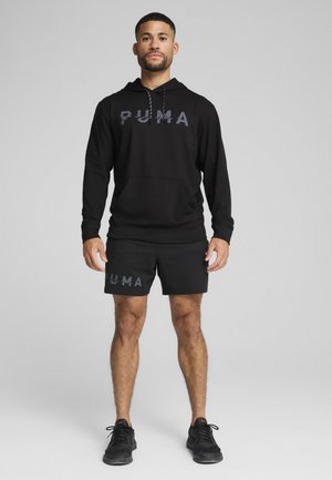Sort hoodie med en frontlomme og snoretræk, der har et stort gråt "PUMA" logo. Stylet med matchende sorte shorts og løbesko.