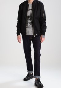 Veste bomber noire avec fermeture éclair frontale et poche latérale, t-shirt gris avec motif tête de mort, jean en denim foncé, et chaussures en cuir noir avec revers retroussés.