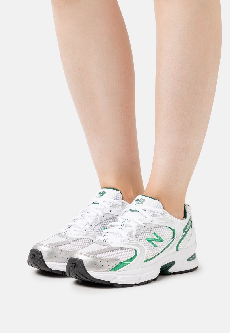 Piernas que llevan zapatillas de running New Balance blancas y verdes con suelas negras, de pie sobre una superficie blanca.