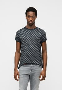 Νέος άνδρας που στέκεται, φοράει μαύρο και γκρι κοντομάνικο πουκάμισο με σχέδιο και ανοιχτό γκρι τζιν, σε απλό λευκό φόντο.