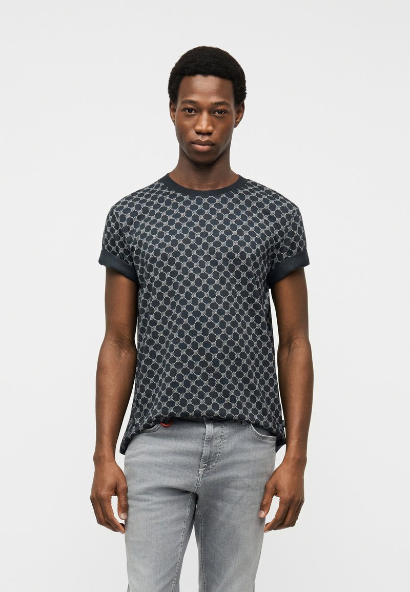 Νέος άνδρας που στέκεται, φοράει μαύρο και γκρι κοντομάνικο πουκάμισο με σχέδιο και ανοιχτό γκρι τζιν, σε απλό λευκό φόντο.