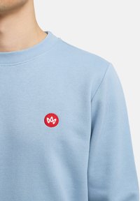 Felpa azzurra chiara con un logo rosso ricamato rotondo sul petto. Presenta un collo a girocollo con costine e un tessuto morbido e texturizzato.