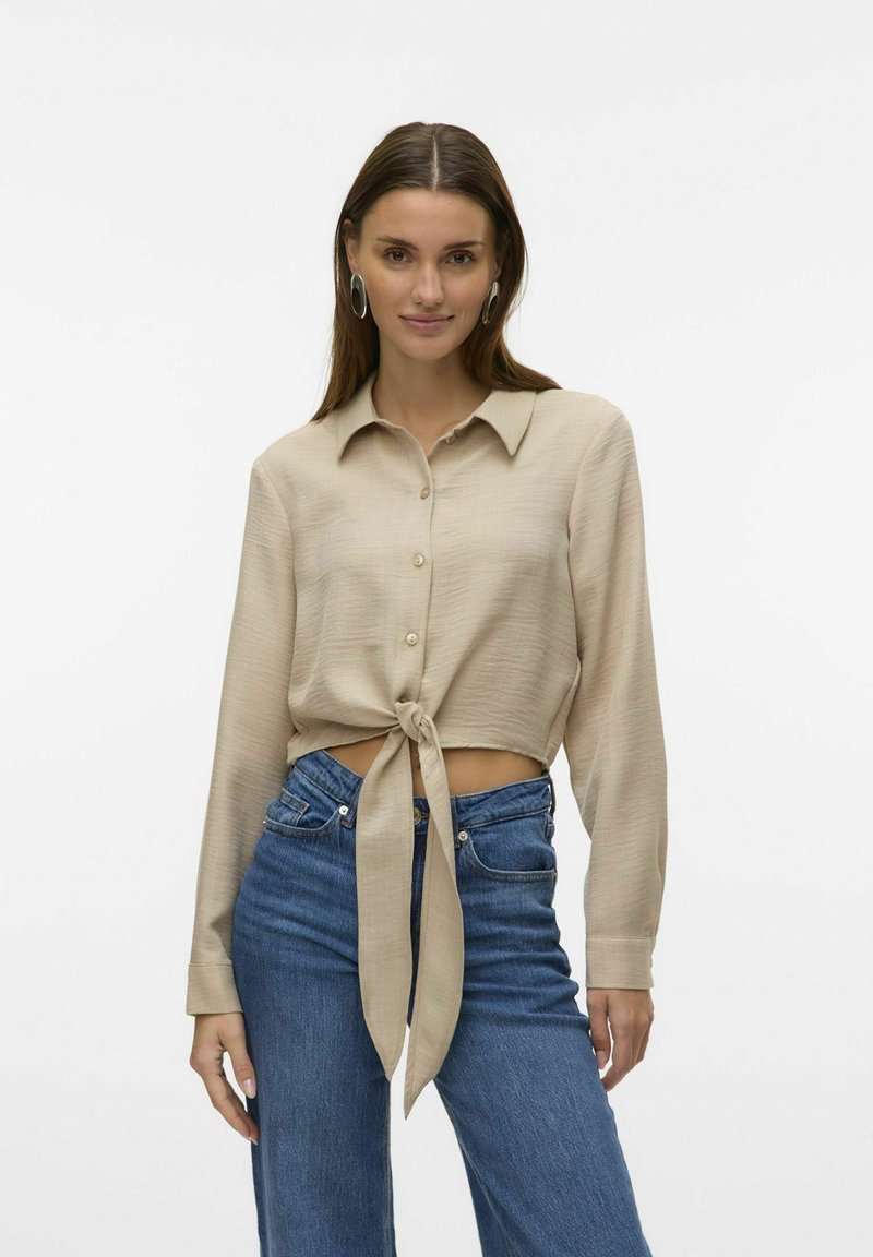 Camicia beige a maniche lunghe con nodo frontale, design con bottoni, texture morbida e colletto classico, indossata con jeans blu a vita alta.