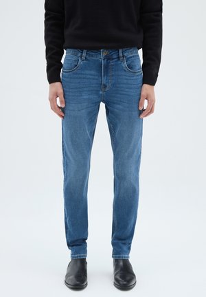 Hombre con jeans ajustados azules, suéter negro y botas de cuero negras hasta el tobillo, de pie contra un fondo claro y sencillo.