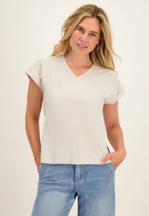Vrouw met lang blond haar draagt een lichtbeige V-hals T-shirt en blauwe jeans, glimlachend met één hand in haar zak tegen een effen achtergrond.