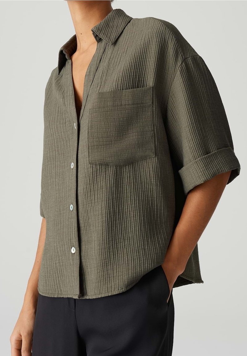 Femme portant une chemise boutonnée texturée vert olive avec une poche avant et des manches retroussées, associée à un pantalon noir, la main dans la poche.