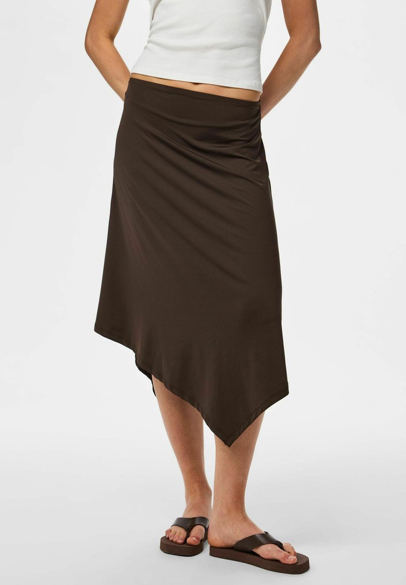 Femme portant une jupe midi asymétrique marron foncé, un top court blanc et des sandales marron foncé, debout, les mains dans le dos.