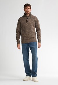 Mężczyzna ubrany w brązowy sweter z guzikami, niebieskie dżinsy i białe trampki, stojący na tle jednolitego białego tła.