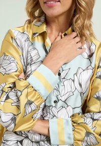Satin blouse features a floral pattern with yellow and blue accents, and a button-down front. Cuffs have a striped yellow detailing.