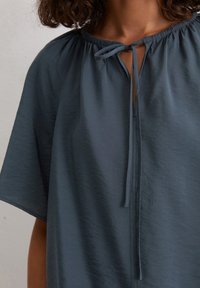 Top comodo di colore blu scuro, con scollatura rotonda, collare arricciato e dettaglio con nodo davanti. Tessuto morbido e testurizzato con maniche corte.