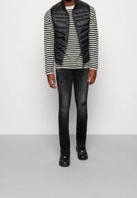 Czarna pikowana kamizelka na białym i czarnym swetrze w paski, zestawiona z ciemnymi dżinsami z przetarciami i czarnymi trampkami.