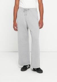 Mand iført lysegrå, vide sweatpants med sn�øre i livet og sorte sneakers med hvide striber, stående mod en hvid baggrund.
