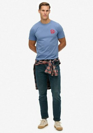 Man staat met zijn handen achter zijn rug, gekleed in een lichtblauw T-shirt, donkere spijkerbroek, beige sneakers en een geruite shirt om zijn taille geknoopt.