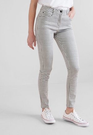 Frau trägt hochtaillierte, schmale Hosen mit grauem und weißem Vichy-Karo-Muster und weiße Sneakers mit niedrigem Schaft, steht mit einer Hand in der Tasche.