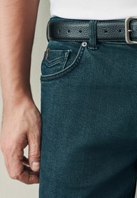 Vaqueros de mezclilla azul oscuro con un cinturón de cuero negro texturizado. Presentan un bolsillo frontal con un distintivo acento de costura dorada.