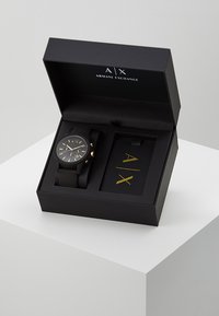 Montre noire avec un cadran rond, des accents dorés et un bracelet en caoutchouc présentée dans une boîte noire arborant le logo doré à l'intérieur.