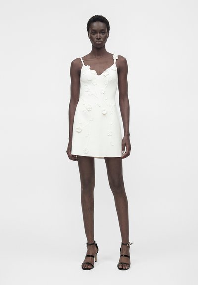 Η γυναίκα στέκεται μπροστά, φορώντας ένα λευκό μίνι φόρεμα χωρίς μανίκια με floral απλικέ και μαύρα ψηλοτάκουνα σανδάλια σε απλό φόντο.