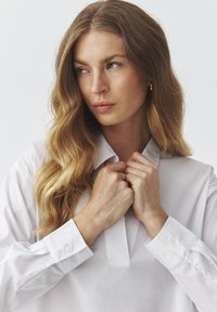 White long-sleeve shirt featuring a placket neck, buttoned cuffs, and smooth texture. Model holds the collar, showcasing the shirt's fit.