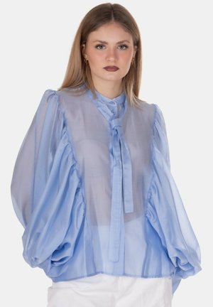 Giovane donna che indossa una camicia azzurro chiaro trasparente con maniche a sbuffo e dettaglio annodato al collo, abbinata a pantaloni bianchi, su uno sfondo neutro.