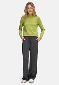 Nő áll a kezét a zsebébe dugva, lime zöld, hosszú ujjú felsőben, laza fekete nadrágban és fehér tornacipőben egy fehér háttér előtt.