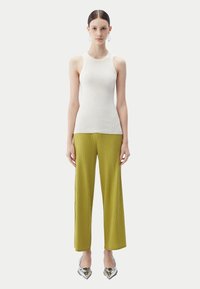 Ung kvinna med bakåtstruket hår som bär vit ärmlös ribbstickad topp, olivgröna vida ribbstickade byxor, silvriga spetsiga skor och ringörhängen.