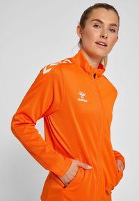 Lys orange zip-jakke lavet af glat stof, med hvidt logo og chevron-detaljer på skuldrene, samt sidelommer og høj krave.