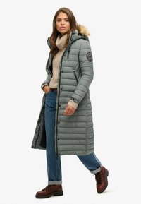 Abrigo largo y acolchado de color verde con capucha con borde de piel, cierre frontal con cremallera, textura acolchada, llevado sobre un suéter beige y jeans azules, combinado con botas marrones.