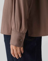 Lange mouwen bruine blouse met een knoopsluiting bij de manchetten, gemaakt van een gladde stof. De blouse heeft een ontspannen pasvorm en wordt gecombineerd met donkere spijkerbroeken.