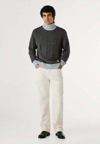 Pullover grigio scuro a coste con colletto alto, abbinato a pantaloni color crema e scarpe nere, creando un look casual.
