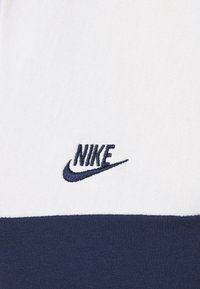 Nike-logotyp broderad på vitt tyg med ett marint horisontellt rand, med en mjuk textur och rena linjer.
